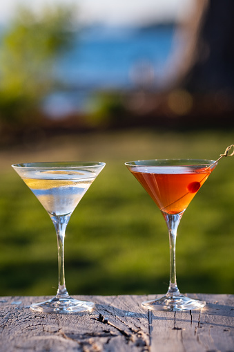 One ice cold Martini with a twist, and one ice cold Manhattan with a cherry sit outdoors on a wood table in a backyard by a lake.