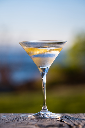 A Martini Cocktail w/ lemon twist sit outdoors on a wood table in a backyard by a lake.