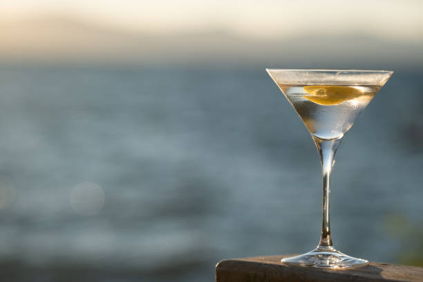 Lakeside Martini at Sunset A Martini Cocktail w/ lemon twist sit outdoors on a wood table in a backyard by a lake. The sun is setting and the light is soft and golden. golden hour drink stock pictures, royalty-free photos & images