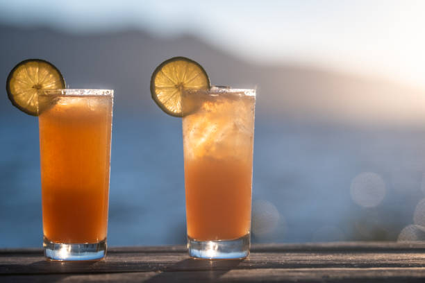 Two orange cocktails on the rocks (could be Mai Tai's, Tequila Sunrises, Screwdrivers, or any number of drinks) garnished with lime wheels sit outdoors on a wood table by a lake. The sun sets in the background illuminating the cocktails.