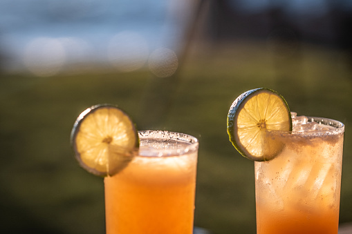 Two orange cocktails on the rocks (could be Mai Tai's, Tequila Sunrises, Screwdrivers, or any number of drinks) garnished with lime wheels sit outdoors on a wood table by a lake. The sun sets in the background illuminating the cocktails.