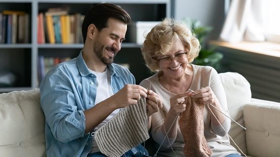 Happy mature mother and son knitting together, enjoying leisure time at home, smiling middle aged woman wearing glasses and young man sitting on cozy couch in living room, holding needles