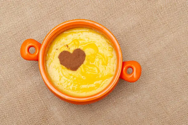 Photo of Homemade sweet corn pudding known in Brazil as Curau or Canjica Nordestina in ceramic bowl. Powdered cinnamon in the heart shape. Isolated on jute background. Copy space. Top view. Horizontal shot