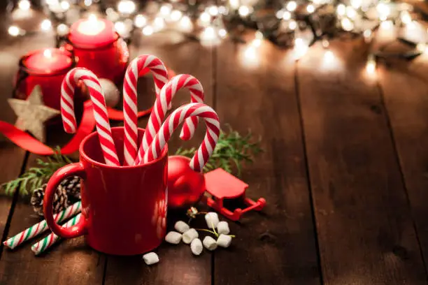 High angle view of a red mug with candy canes inside shot on rustic wooden table. The mug is surrounded by some Christmas ornaments. Christmas lights are out of focus at background. The composition is at the left of an horizontal frame leaving useful copy space for text and/or logo. Predominant colors are red and brown. Low key DSRL studio photo taken with Canon EOS 5D Mk II and Canon EF 100mm f/2.8L Macro IS USM.