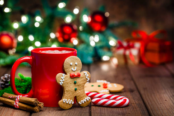 Gingerbread cookie and hot chocolate for Christmas Christmas backgrounds: gingerbread cookies and hot chocolate shot on rustic wooden table. The composition is at the left of an horizontal frame leaving useful copy space for text and/or logo at the right. Christmas tree and string lights are out of focus at background.  Predominant colors are red, green and brown. traditional christmas stock pictures, royalty-free photos & images
