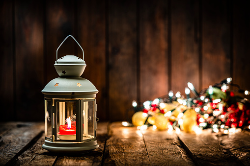 Christmas backgrounds: Christmas lantern on rustic wooden table. The composition is at the left of an horizontal frame leaving useful copy space for text and/or logo at the right. String light and Christmas decoration are out of focus at background. Predominant colors are yellow and brown. High resolution 42Mp studio digital capture taken with Sony A7rII and Sony FE 90mm f2.8 macro G OSS lens