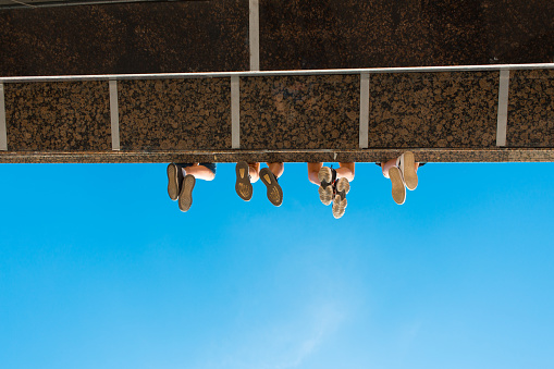 Boys legs in shoes hanging from the bridge against blue sky.