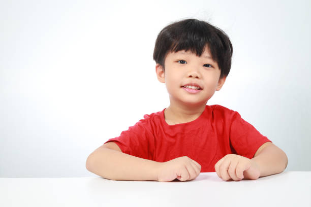Portrait of a cheerful little Asian boy sitting at the desk, looking at camera, isolated on white background