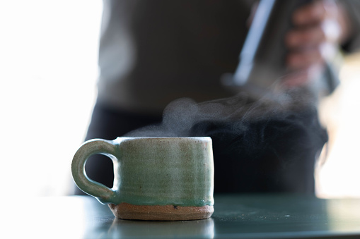 close up of steam drifting from natural ceramic coffee cup with blurred barista in background