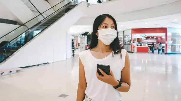 New normal shoppping lifestyle concept, Traveler asian woman with mask to protect coronavirus outbreak using mobile phone in shopping mall at bangkok, Thailand