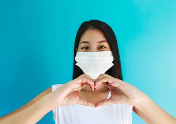 Happy asian woman with medical mask making hand on heart shape in blue screen background, Coronavirus or Covid-19 new normal and social distancing concept