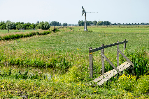 Hiking trail through the polder near Schipluiden, the Netherlands with a narrow wooden bridge, a steel windmill and in the distance a church tower