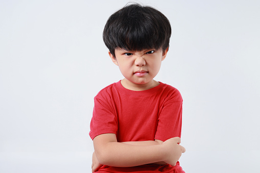 Portrait of frustrated little Asian boy with red t-shirt, arms crossed, showing angry face, looking at the camera over white background.