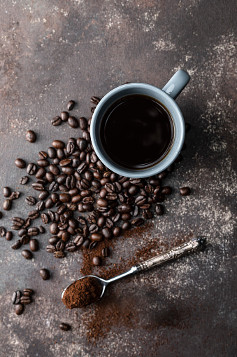 Direct above view of roasted coffee beans, silver spoon and blue coffee cup with black coffee on dark brown background.