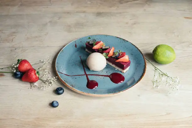 landscape shot of a plated strawberry slice dessert with sorbet, on a blue plate and pale wood backdrop, with elderflower, lime, strawberries and blueberries