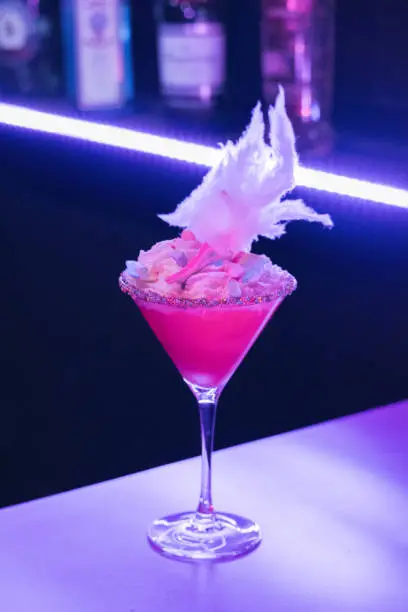 vertical image with blured background of a single pink cocktail with sugared rim and candy floss in a martini glass, bar bottles visible in the background