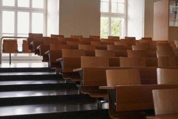Empty auditorium at university with wooden chairs and banks and large windows and stairs on one side. Empty auditorium at university with wooden chairs and banks and large windows and stairs on one side. The chairs are arranged in rows and their backrests form a pattern. lecture hall stock pictures, royalty-free photos & images