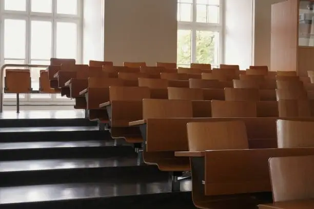 Photo of Empty auditorium at university with wooden chairs and banks and large windows and stairs on one side.