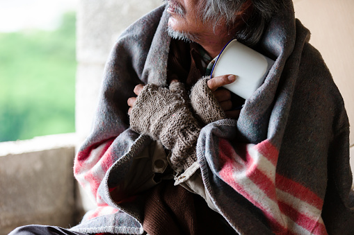 Poor old homeless asian man sitting with dirty blanket, gloves sitting cold in the corner of an abandoned building.
