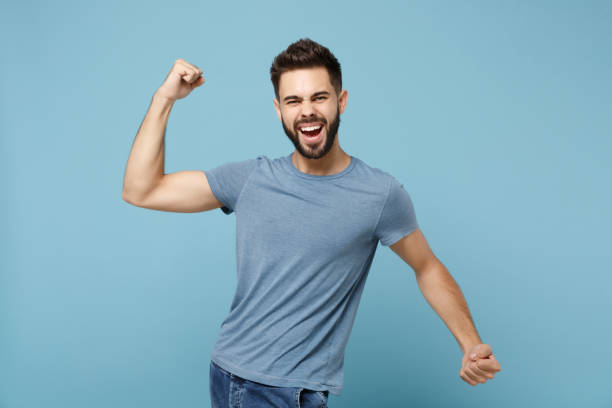 istock Young laughing handsome man in casual clothes posing isolated on blue wall background, studio portrait. People sincere emotions lifestyle concept. Mock up copy space. Clenching fists like winner. 1270987192