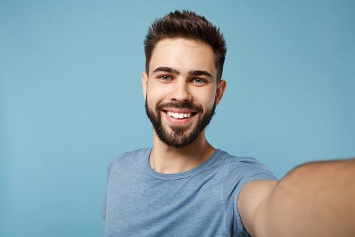 Close up Young smiling man in casual clothes posing isolated on blue wall background, studio portrait. People sincere emotions lifestyle concept. Mock up copy space. Doing selfie shot on mobile phone.