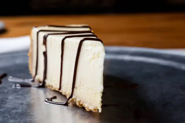 A closeup view of a slice of New York cheesecake with chocolate syrup, in a restaurant or kitchen setting.