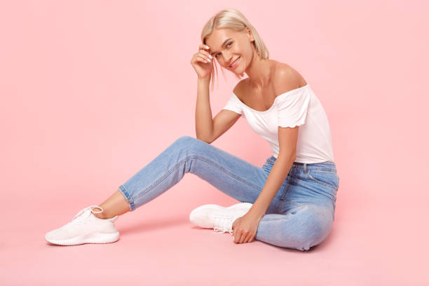 Young adult woman in denim wear clothes looking at camera with smile Full length view of cheerful and smiling young adult woman sitting on pastel pink background, looking at camera. Happy girl posing in casual denim wear and white sneakers girl sitting stock pictures, royalty-free photos & images