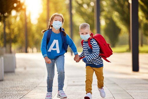 Delighted little sister and brother in casual clothes and protective masks with backpacks holding hands and running together in park ready for school studies