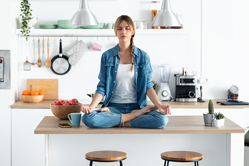 Shot of pretty young woman in lotus position sitting on the table while relaxing in the kitchen at home.