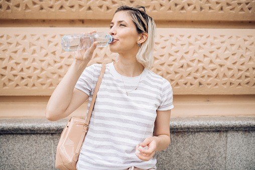 Cheerful young woman with a blonde hair, drinking water from a plastic bottle on a hot day, hydrating