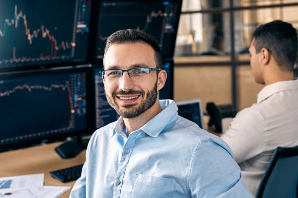istock Teamwork Concept. Traders sitting at office together monitoring stocks data on screen bearded man looking back camera cheerful achieving success close-up 1271031315