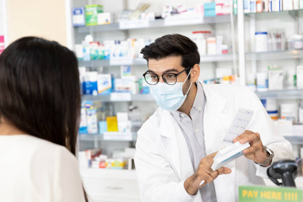 Pharmacist wearing protective hygienic mask and making drug recommendations in modern pharmacy Middle eastern male pharmacist wearing protective hygienic mask to prevent infection selling medications to woman patient to prescription and making drug recommendations in modern pharmacy chemist photos stock pictures, royalty-free photos & images