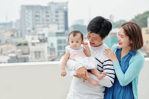 Happy family of young Asian father, mother and little daughter standing on balcony of their apartment