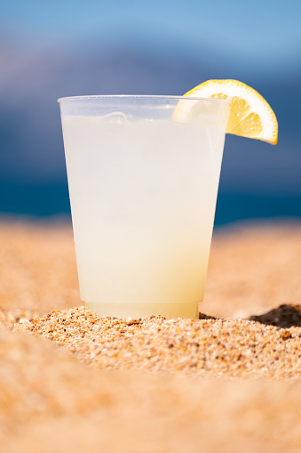 The perfect glass of lemonade, garnished with a slice of fresh lemon, rests on the golden sandy beach of a bright blue mountain lake. The perfect summer beverage in the perfect summer setting.