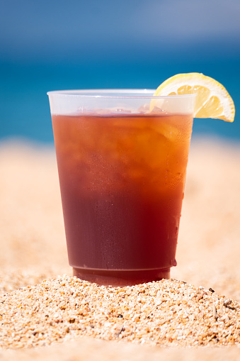 The perfect glass of iced tea, garnished with a slice of fresh lemon, rests on the golden sandy beach of a bright blue mountain lake. The perfect summer beverage in the perfect summer setting.