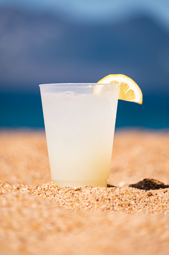 The perfect glass of lemonade, garnished with a slice of fresh lemon, rests on the golden sandy beach of a bright blue mountain lake. The perfect summer beverage in the perfect summer setting.