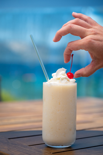 A creamy frozen white Pina Colada Cocktail sits on a beach side wooden table. A hand carefully sets a cherry on top of a whipped cream garnish. Tropical blues and greens in the background.
