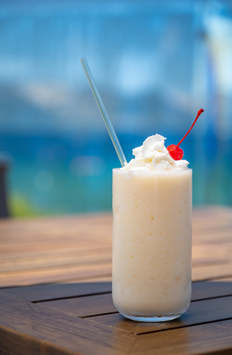 A creamy frozen white Pina Colada Cocktail sits on a beach side wooden table. A cherry sits perched atop a whipped cream garnish. Tropical blues and greens in the background.