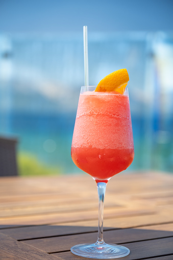 A frozen red cocktail (could be a mai tai, or a hurricane, a rum runner or a strawberry daiquiri) garnished with an orange slice sits on a lake side wooden table. The vibrant greens and blues of the water in the background.