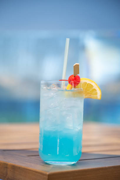 Blue Cocktail with Garnish on a Summer Day A refreshing blue cocktail on the rocks (Could be an AMF or a Blue Motorcycle or a Blue Hawaii) garnished with a lemon and a cherry sits lake side on a wooden table. The vibrant blues and greens of the lake in the background. blue hawaiian stock pictures, royalty-free photos & images