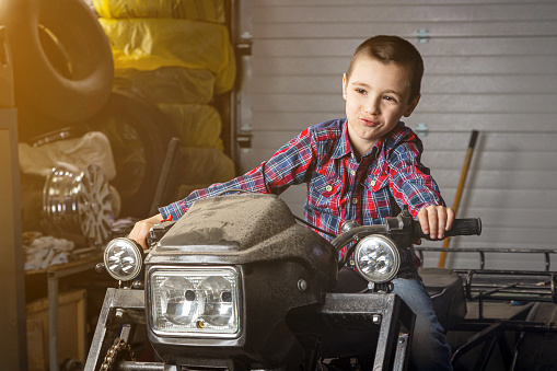 Little boy young auto mechanist cheerfully dreams that he
  rides fast on a motorcycle in the garage of a service station. A child sits on an old ATV