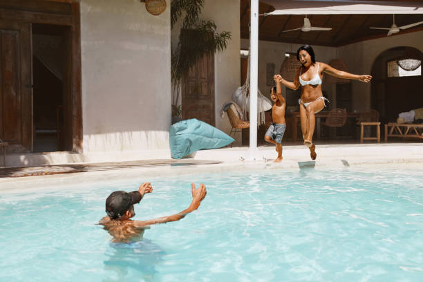 プール。プールサイドから水に飛び込む母と息子。父は子供とお母さんを捕まえる準備をしています。トロピカルリゾートで休暇を楽しむ家族。ライフスタイルとしての夏の活動。 - men child swimming pool women ストックフォトと画像