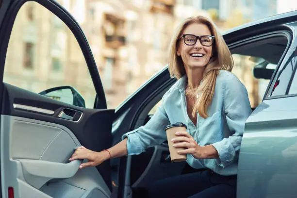 Happy attractive woman or business lady wearing eyeglasses holding cup of coffee and getting out of her modern car, transportation concept