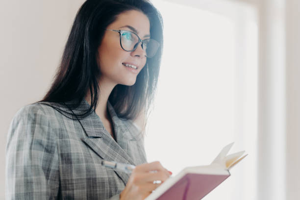 istock Serious thoughtful brunette European woman writes down text information in notepad, concentrated somewhere into distance, makes notes, wears formal clothes, transparent glasses, stands indoor 1271094058