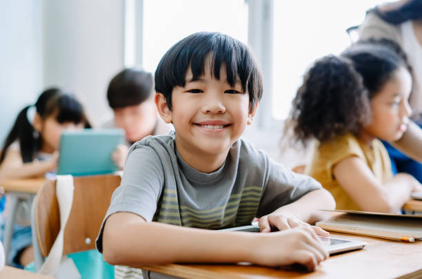 cute little asian boy uśmiechając się patrząc na kamerę i za pomocą laptopa w klasie komputera w szkole podstawowej. edukacja, szkoła, technologia i internet. - korea child baby asian culture zdjęcia i obrazy z banku zdjęć