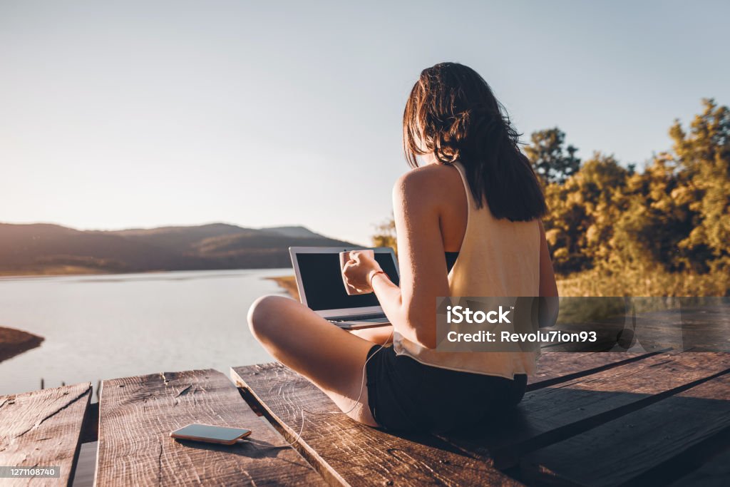 Young Woman using laptop outdoor on wooden lake pier Woman working on laptop in the nature, she sitting on wooden pier around beautiful mountain lake and using laptop in the morning Working Stock Photo