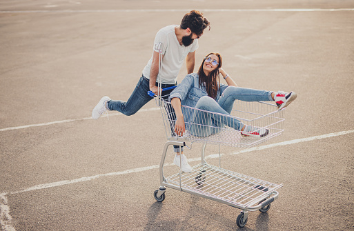 Full body crazy bearded man and cheerful woman looking at each other while riding shopping cart on asphalt parking lot