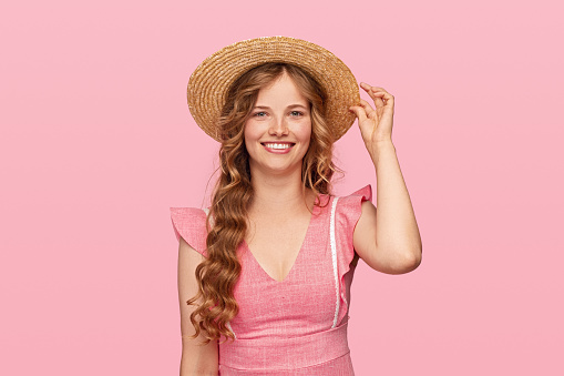 Happy young Russian female smiling for camera and adjusting summer hat against pink background