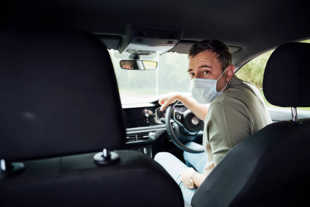 Happy To be Your Driver A shot of a caucasian male in his 40's sat in the driving seat of a car wearing a face mask and looking over his shoulder at camera. taxi driver photos stock pictures, royalty-free photos & images