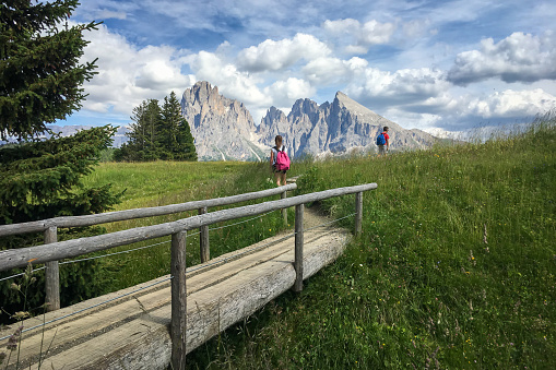 Compatsch, Italy - July 9, 2018: Alpe di Siusi - Seiser Alm with Sassolungo - Langkofel mountain group in front of blue sky with clouds.  Wooden footbrigde with two children hiking during summer in ski resort, Dolomites, Trentino Alto Adige, South Tyrol, Italy
